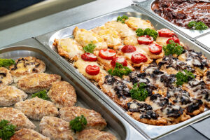Various dishes displayed in a catering service setting. The left tray contains breaded patties, while the right tray features chicken topped with cheese, tomatoes, and mushrooms. Fresh parsley garnishes the dishes. Catering Riesa.