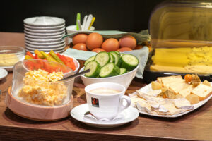 A breakfast display with scrambled eggs in a wooden bowl, slices of cucumber, tomatoes, boiled eggs, and assorted cheeses. A cup of coffee is on a white saucer, with stacks of plates in the background. The setting suggests a catering arrangement for a morning meal.