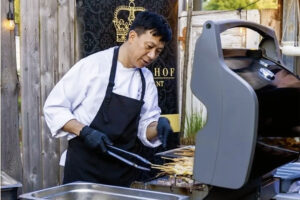 A chef in an apron and gloves cooking skewers on a grill outdoors, focusing on the food. In the background, a sign with a crown logo is visible, suggesting a culinary establishment, possibly related to Catering Riesa.